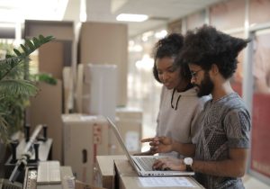 Side view of young African American female and ethnic man wearing casual clothes working on project using laptop while standing near boxes